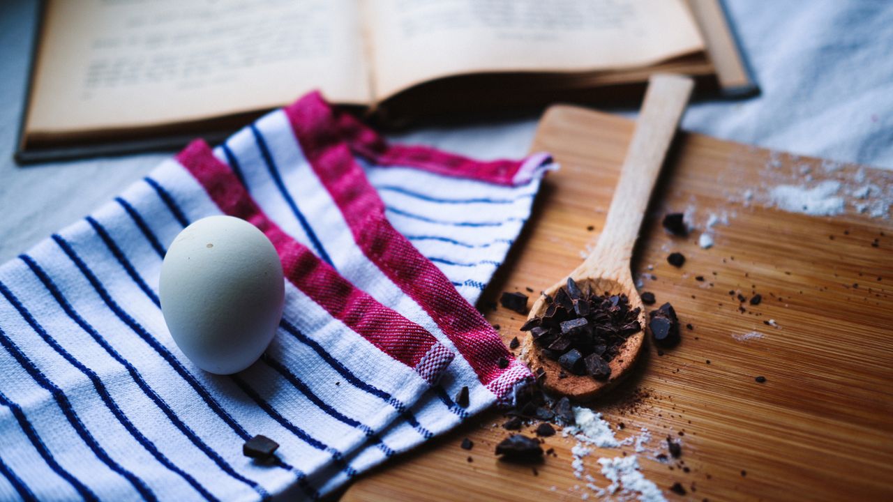 hero-gallery-03 Overhead view of baking essentials with egg, chocolate, and open cookbook on wooden table.
