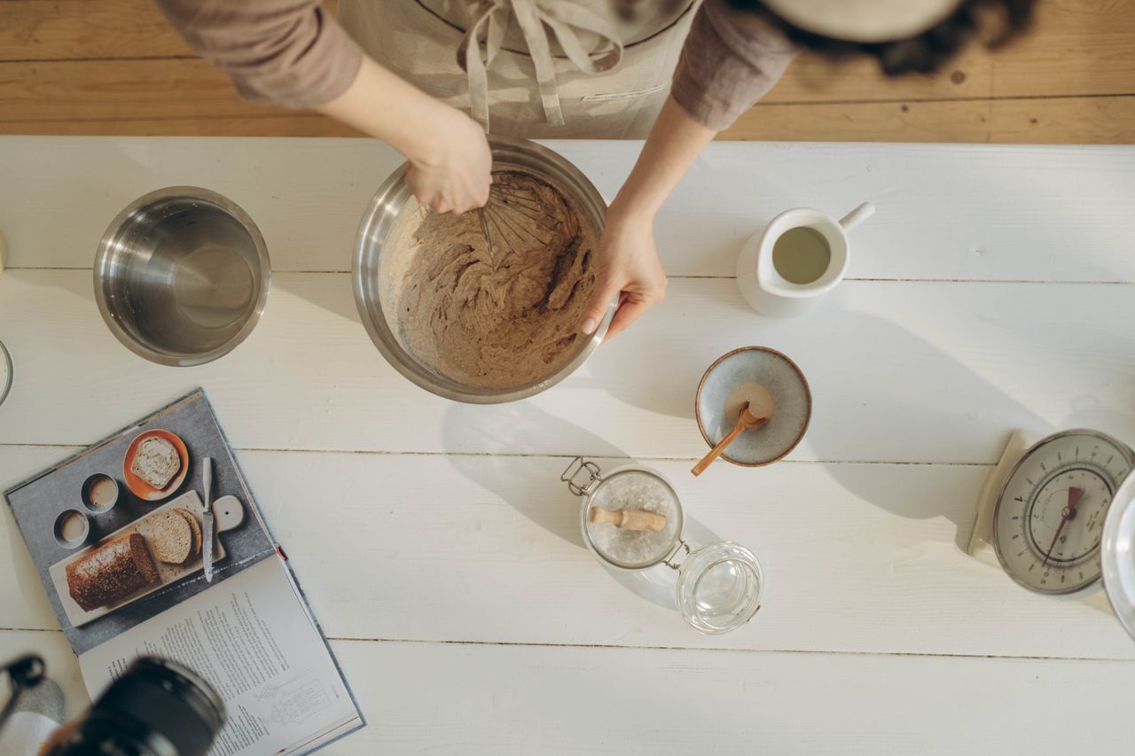 hero-gallery-01 A close-up of hands mixing batter in a bowl among baking ingredients on a wooden table.